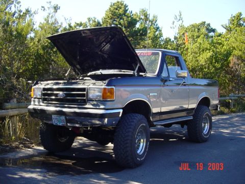 1989 Ford Bronco - A two-tone gray and black Ford Bronco sits with its hood open, modified with a lifted suspension and large off-road tires.