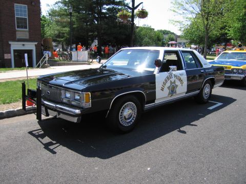 1991 Ford LTD Crown Victoria - black-and-white Ford Highway Patrol sedan with a front push bar parked on pavement near a brick building.