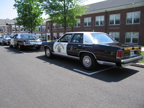 1991 Ford LTD Crown Victoria - Rear three-quarter view of a black-and-white Highway Patrol sedan parked in a marked lot with other patrol cars.