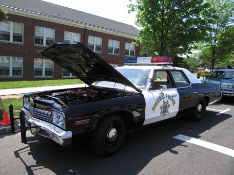 1991 Ford LTD Crown Victoria - black-and-white Highway Patrol sedan with raised hood revealing a red engine, parked near a brick building.