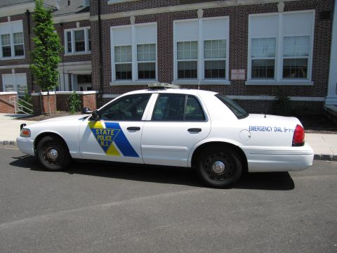 1991 Ford LTD Crown Victoria - white New Jersey State Police sedan marked 'Emergency Dial 9-1-1' parked along a street by a brick building.