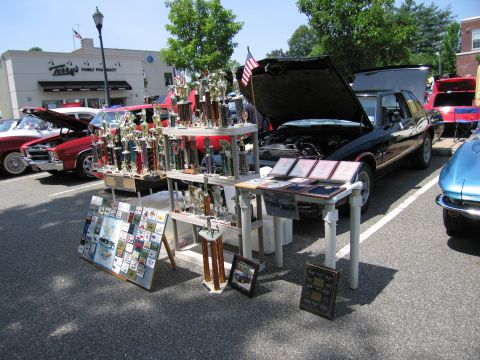 1991 Ford LTD Crown Victoria - table of trophies and plaques set up beside a black car with an open hood at an outdoor car show.