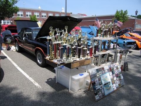 1991 Ford LTD Crown Victoria - Rows of tall trophies and plaques displayed on tables behind a black car with an open hood at a car show.
