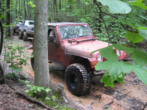1991 Ford Bronco - A red Jeep Wrangler tackles a challenging muddy off-road course, followed by other vehicles.
