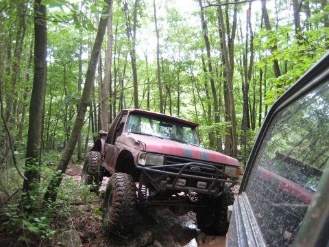 1991 Ford Bronco - A red off-road truck drives through a muddy forest path, with another vehicle partially visible on the right.