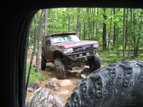 1991 Ford Bronco - A red 1991 Ford Bronco navigates a rocky, muddy trail through a forest.