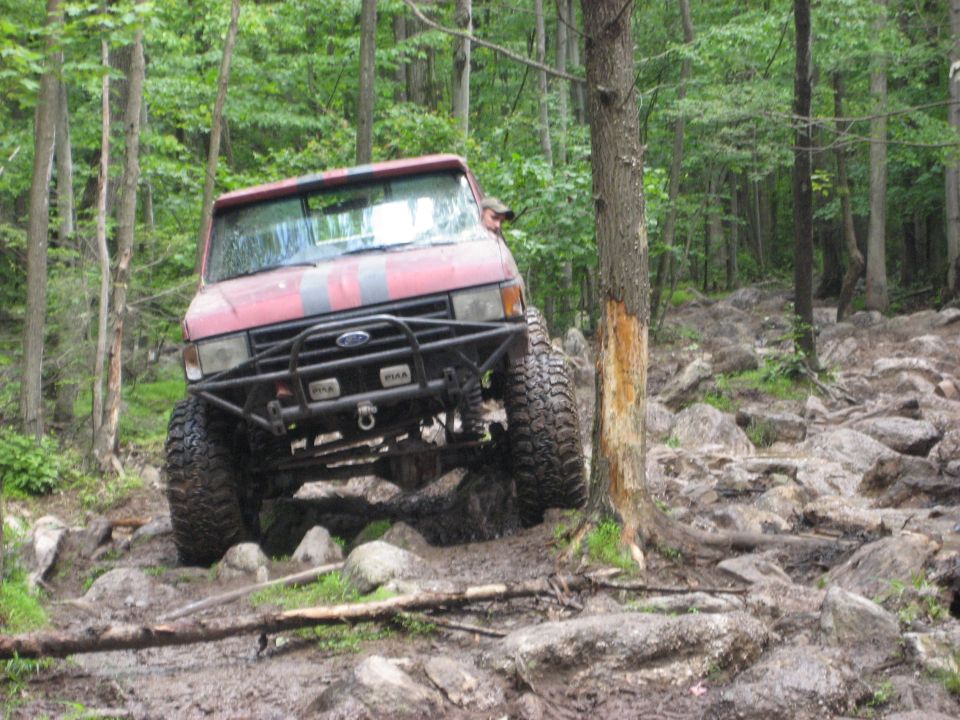 Red off-road SUV with large tires and a brush guard navigating a rocky, muddy trail