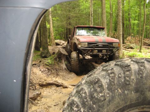 1991 Ford Bronco - A red 1991 Ford Bronco navigates a challenging muddy trail in a wooded area, with another vehicle's tire visible in the foreground.
