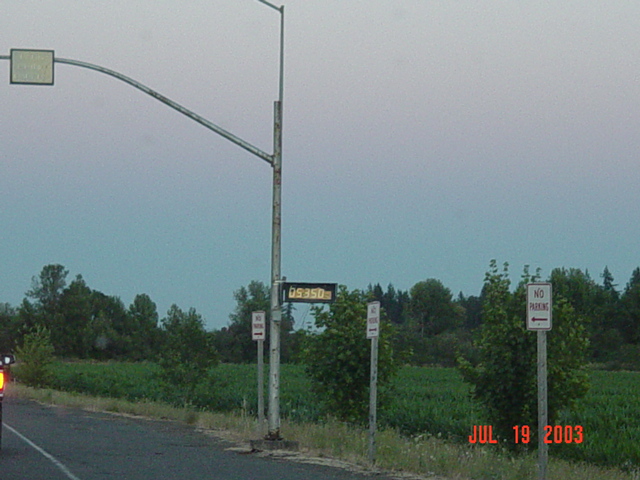 Roadside pole with small digital display and No Parking signs at dusk beside a field