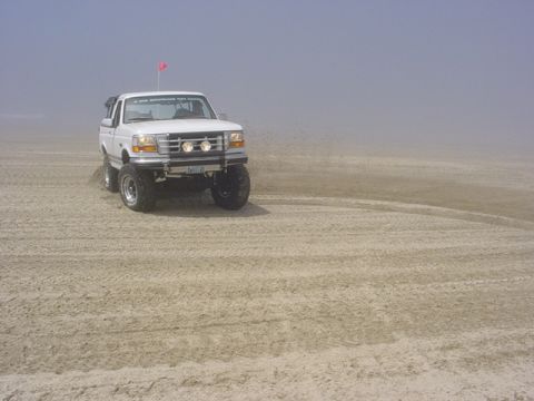 1989 Ford Bronco - This lifted pickup truck is driving on a sandy beach with a hazy sky in the background.