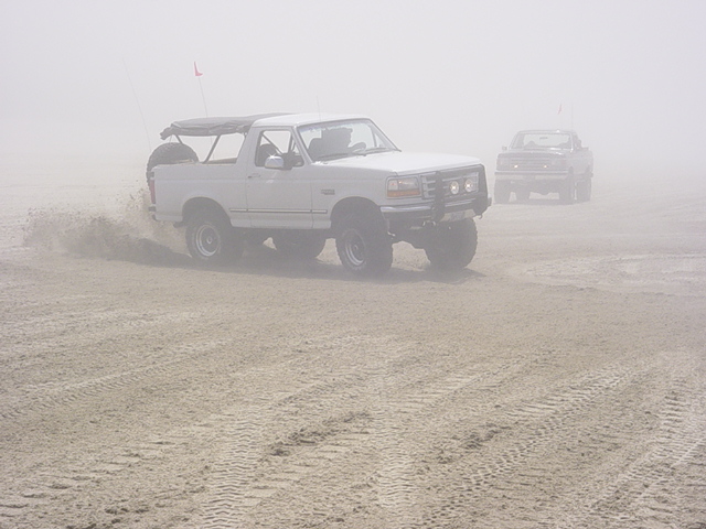 White topless Ford Bronco with brush guard driving across foggy beach sand, spraying sand