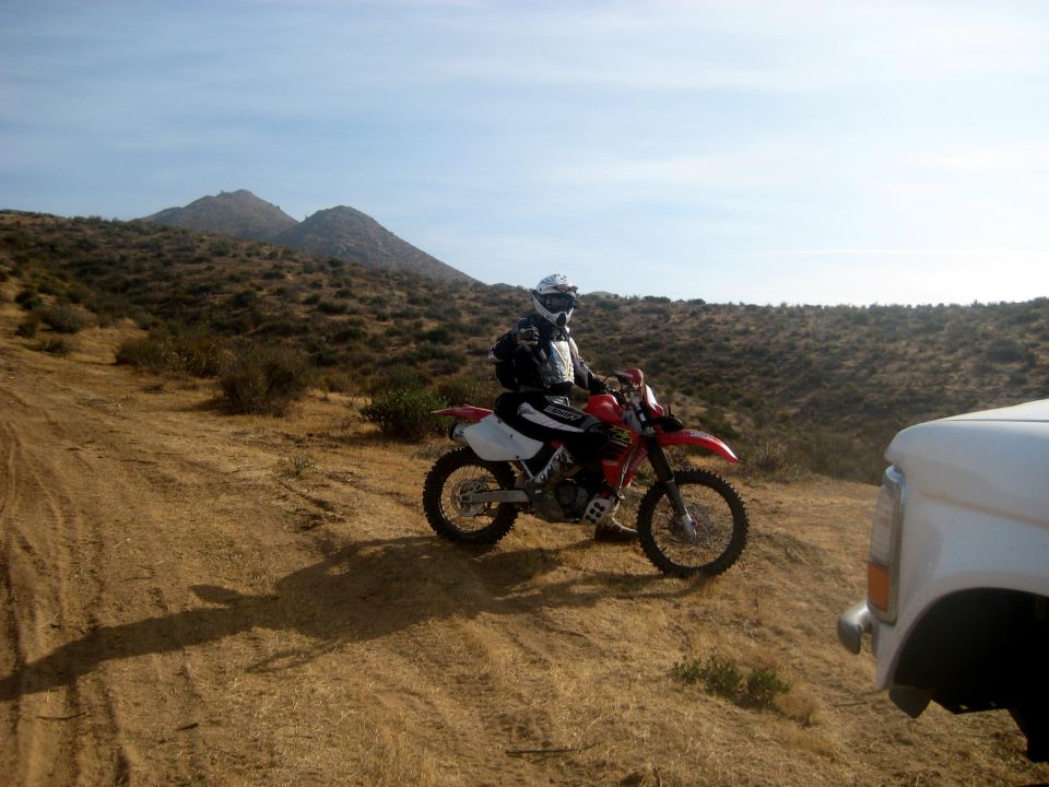 Dirt bike rider in full gear sits on a red dirt bike on a dry, grassy hill. Part of a white truck is visible on the right
