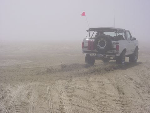 1989 Ford Bronco - A white 1989 Ford Bronco drives on a sandy beach, kicking up dust in the fog.