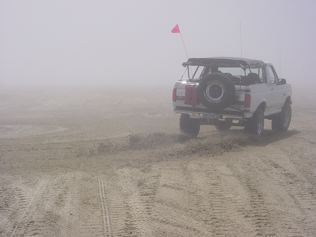White Ford Bronco with spare tire carrier and pink dune flag on foggy sand dunes