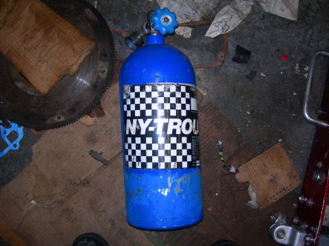 1984 Ford Bronco - Nitrous oxide bottle with checkered flag label, situated near a flywheel and other garage items.