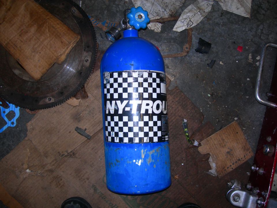 Blue nitrous oxide bottle with checkered flag label reading NY-TROU, next to a flywheel
