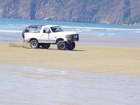 1989 Ford Bronco - A white 1989 Ford Bronco is driven on a sandy beach near the ocean, kicking up sand.