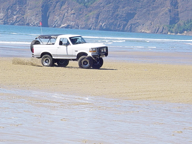White lifted Ford Bronco driving on beach sand near ocean cliffs.
