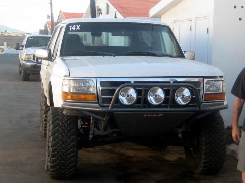 1996 Ford Bronco - A white 1996 Ford Bronco is seen from the front, equipped with an aftermarket bumper and auxiliary lights, with another vehicle in the back…