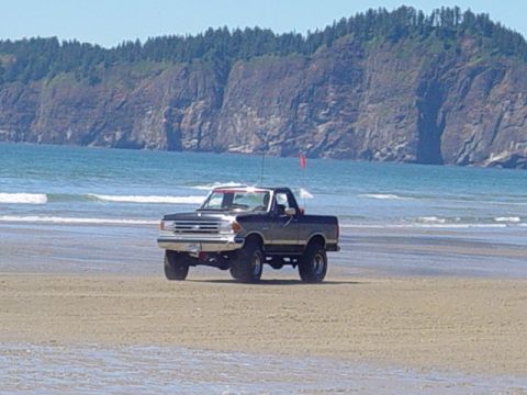 1989 Ford Bronco - A lifted Ford pickup truck is parked on the sand near the ocean waves, with a forested cliff face behind it.