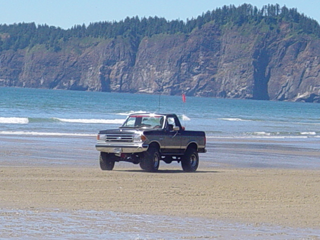 Lifted dark Ford 4x4 with white roof and dune flag driving on an ocean beach