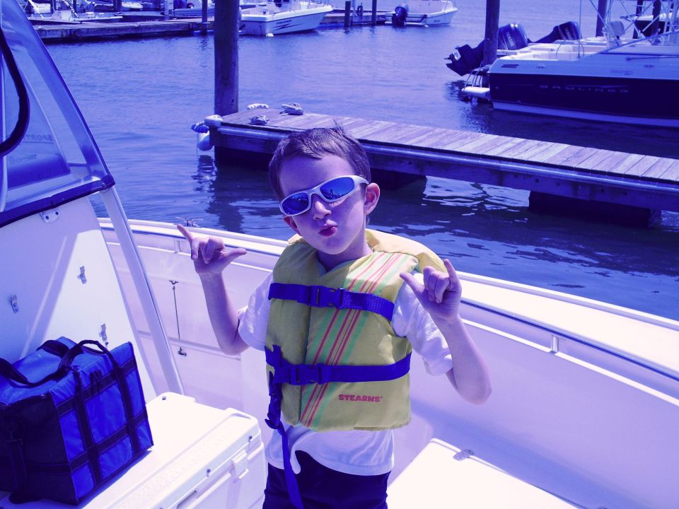 Kid wearing sunglasses and a life jacket on a boat, making 'shaka' hand signs. Other boats and a dock are visible in the background