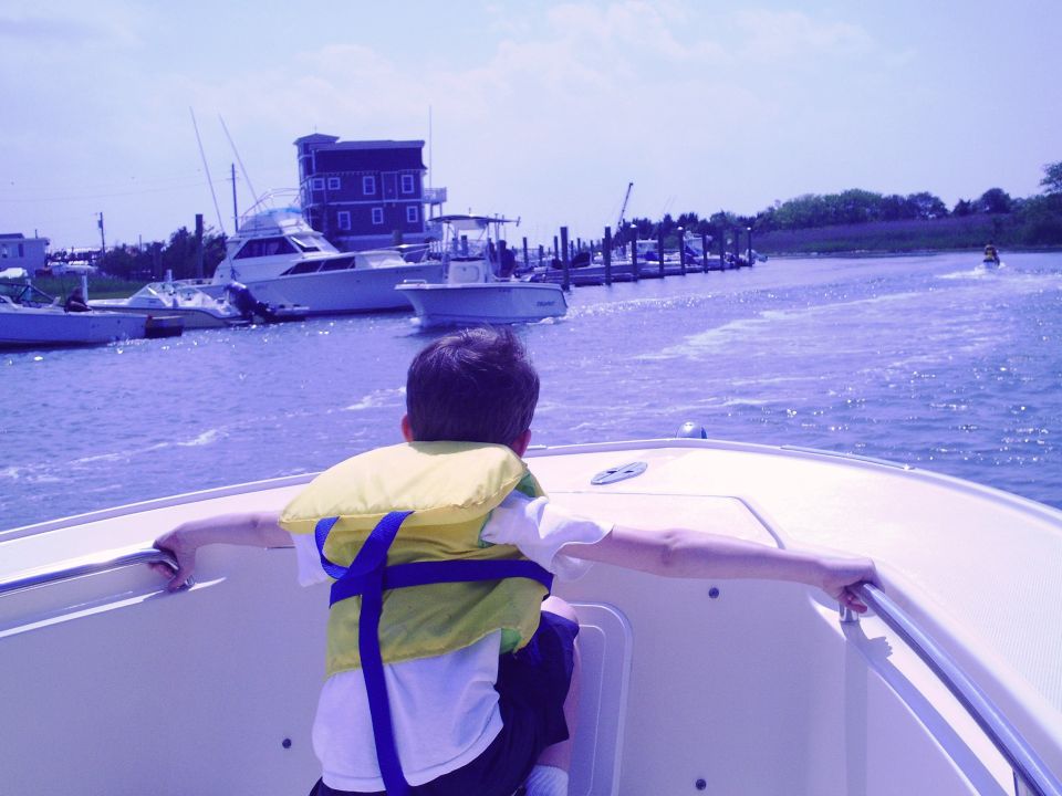 Boy in yellow life jacket on a boat, looking at other boats and a building