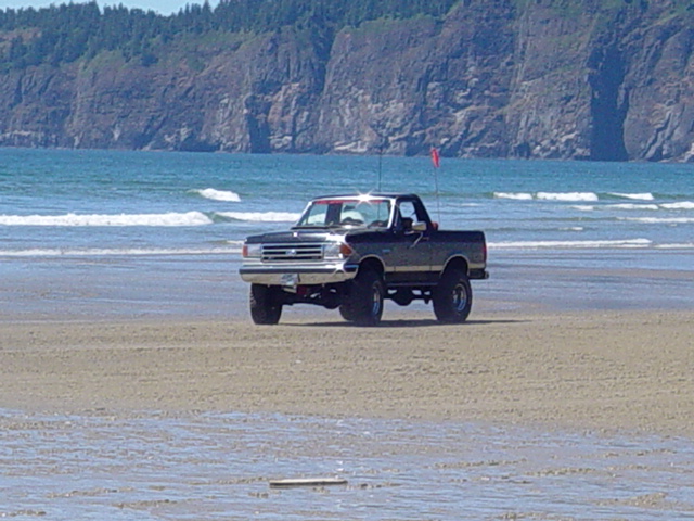 Lifted dark Ford Bronco with top off and dune flag parked on a beach near the surf