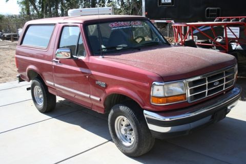 1989 Ford Bronco - This red Ford Bronco is parked on a concrete surface with a trailer and a buggy in the background.