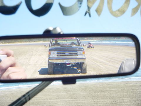 1989 Ford Bronco - Rearview mirror shows a lifted Ford F-Series truck on the beach, with an ATV further down the shore.