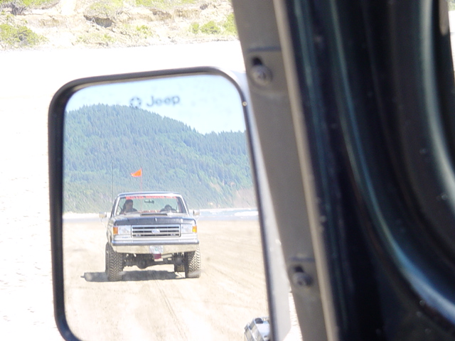 Jeep side mirror reflecting a lifted Ford truck with orange dune flag on sand dunes