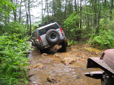 1991 Ford Bronco - A silver SUV tackles a challenging off-road course, splashing through a muddy stream surrounded by lush forest greenery.