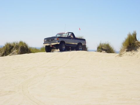 1989 Ford Bronco - This dark blue and silver SUV, likely a 1987 Ford Bronco, navigates a sandy terrain under a clear blue sky.