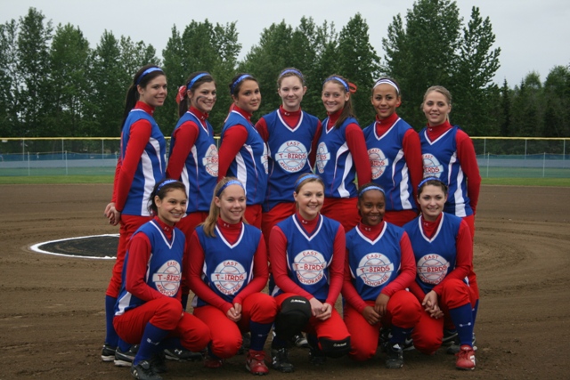 Girls in red and blue softball uniforms pose for a team photo on a baseball field