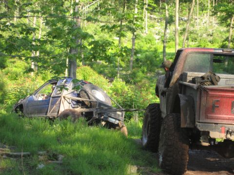 1991 Ford Bronco - A red pickup truck tows a heavily modified, damaged vehicle through a grassy, wooded area.