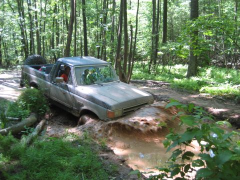 1991 Ford Bronco - A silver pickup truck navigates a muddy trail, splashing water as it gets stuck.