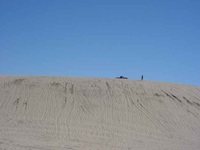 Sand dune covered in tire tracks with a distant truck and person at the crest under blue sky