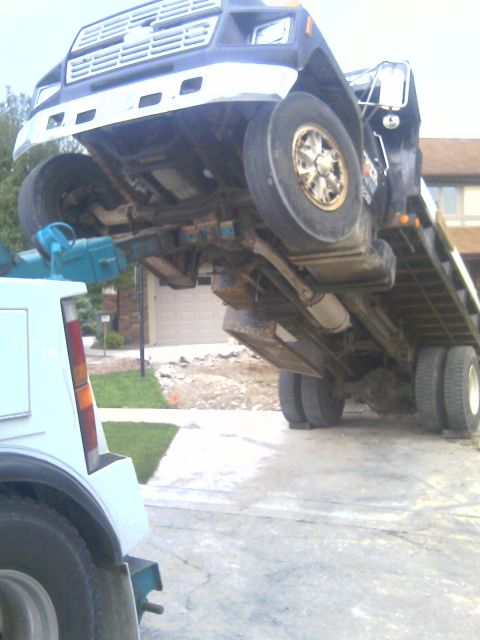 1992 Ford Bronco - A tow truck is lifting the chassis of a blue truck upside down in a driveway.