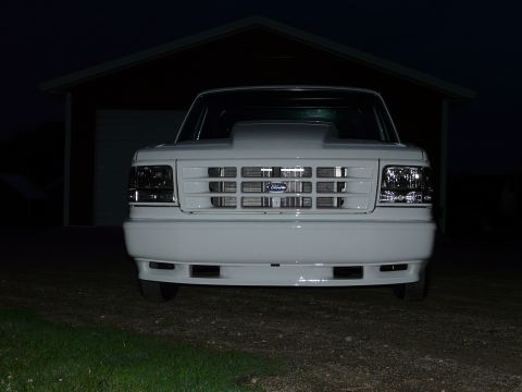 1994 Ford F-150 - Head-on night shot of a white Ford pickup parked on gravel in front of an open pole shed, its body-color grille surround framing a chrome-s…