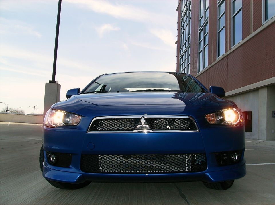 Blue Mitsubishi Lancer photographed head-on with headlights on atop a parking garage