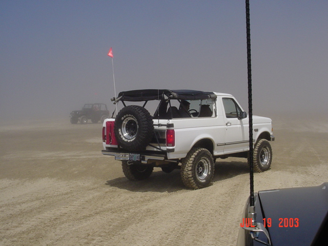 White lifted Ford Bronco with rear tire carrier and whip flag on foggy dunes