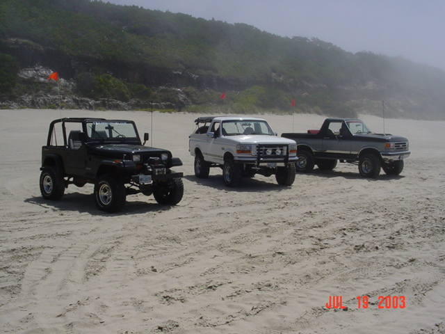 Black Jeep Wrangler, white Ford Bronco, and gray Ford pickup on a beach