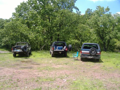 1988 Ford Bronco - Three 1988 Ford Broncos are parked in a grassy area with trees. The middle one has its tailgate open with gear inside.