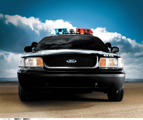 1998 Ford Crown Victoria - A black police car sits on a dirt road under a cloudy blue sky, its emergency lights illuminated.