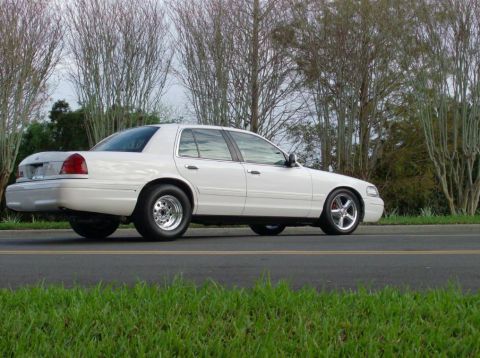1998 Ford Crown Victoria - A white 1998 Ford Crown Victoria sedan is parked on the side of a road with trees in the background.