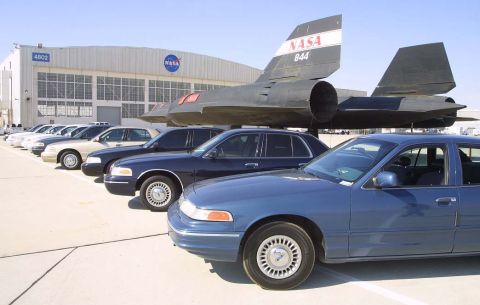 1998 Ford Crown Victoria - A line of Ford Crown Victoria police cars are parked outside a NASA facility, with an SR-71 Blackbird aircraft displayed overhead.