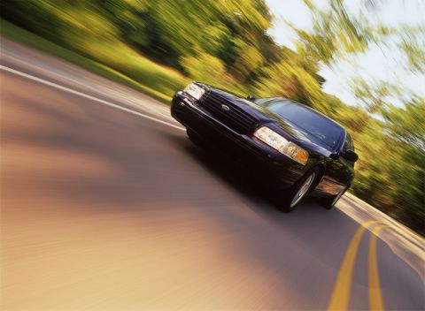 1998 Ford Crown Victoria - A black 1998 Ford Crown Victoria is captured in motion on a two-lane road, with trees lining the side.