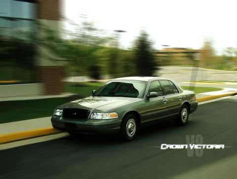 1998 Ford Crown Victoria - Rolling three-quarter front shot of a light green Ford Crown Victoria sedan driving past a brick building, with motion-blurred background,…