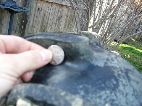 2008 Bombardier Outlander - A hand holds a quarter against a dark, textured vehicle component, with a wooden fence and bare branches in the background.