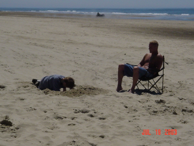 Man sitting in a chair on the beach while a child digs in the sand